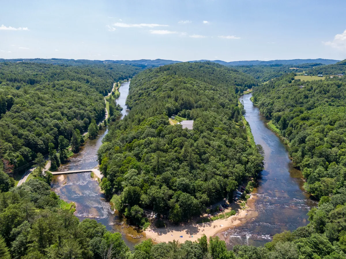 A wide, tranquil river winds through lush green hills, with a sandy beach in the foreground and a bridge crossing over the water. The scene captures the inviting landscape perfect for swimming, kayaking, hiking, and relaxing by the water. The soft light creates a peaceful atmosphere, ideal for outdoor activities.