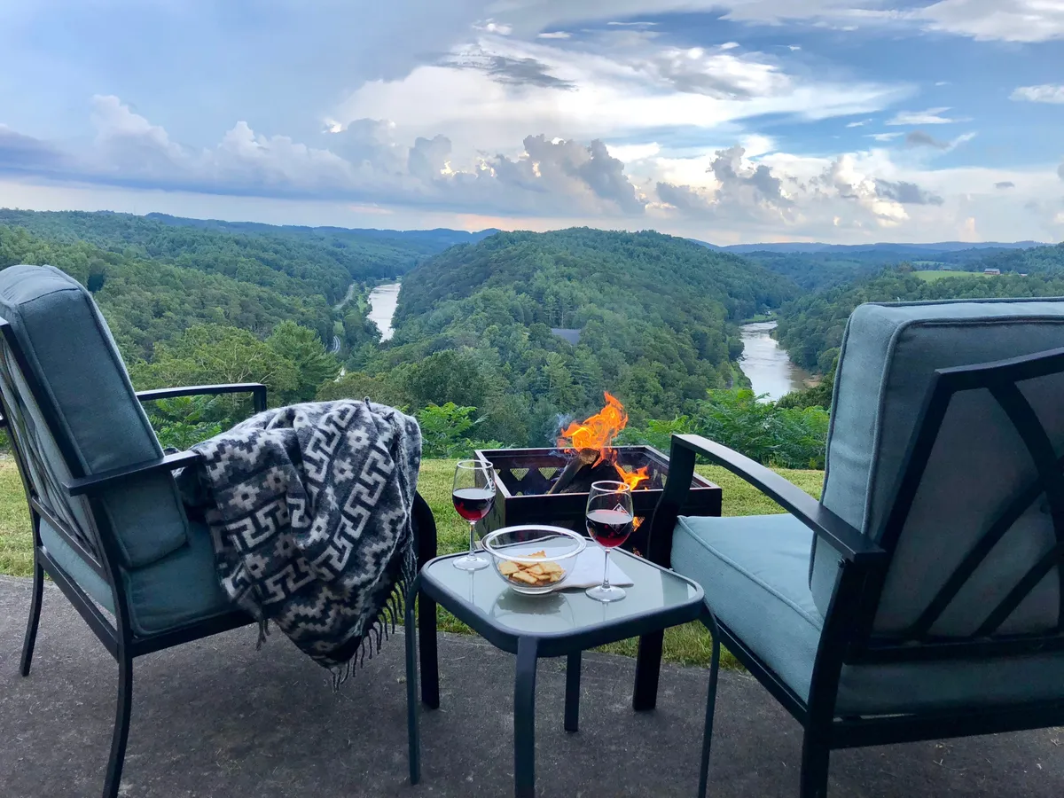 An inviting exterior seating area featuring two comfortable chairs positioned around a fire pit, with a table set for wine and snacks. The backdrop includes breathtaking views of the landscape, suggesting the perfect spot for an intimate evening by the fire, sharing drinks, and taking in the sunset.