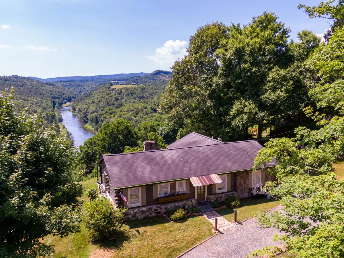 The image showcases a beautifully landscaped single-family house perched above the New River, with a lush garden in the foreground and sweeping, high-elevation river views beyond. The setting glows with the warm hues of sunset, inviting guests to relax outside or enjoy the peaceful natural surroundings. The riverbank is visible, hinting at opportunities for walks, kayaking, or simply unwinding by the water.
