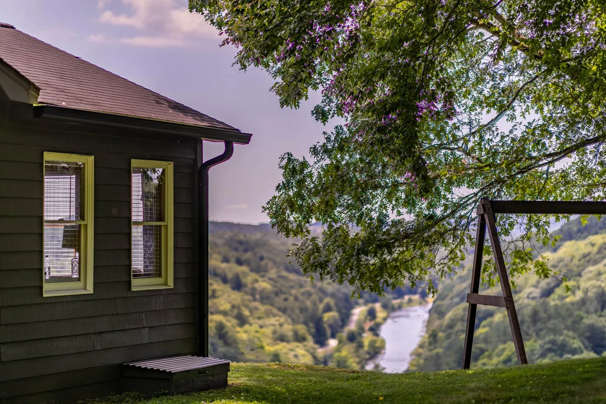 This image showcases the house in its natural setting with a river in the background, a swing set under a large tree, and open grassy space ideal for outdoor activities. The scene exudes tranquility while hinting at the fun and relaxation possible for families, with space for children to play and adults to unwind as they enjoy the views.