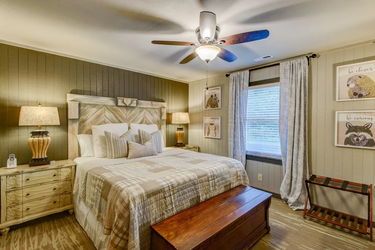 A serene bedroom featuring a plush bed with crisp linens, flanked by two nightstands and softly lit by natural light streaming through curtained windows. A bench sits at the foot of the bed, and tasteful artwork adorns the wall, creating a restful and inviting atmosphere.