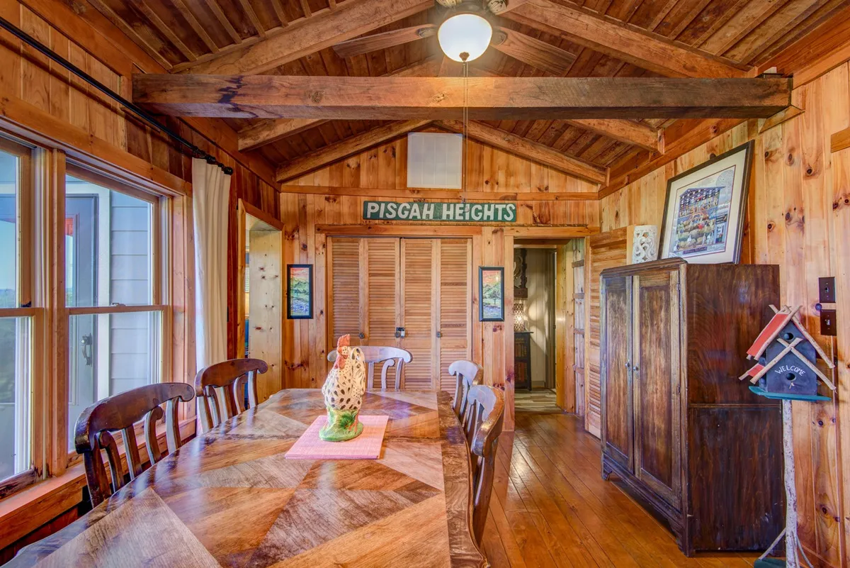 A welcoming dining area with a wooden table set beneath exposed beams, decorative items atop the table, and a 'Pisgah Heights' sign creating a sense of place. The large windows allow sunlight to flood the space.