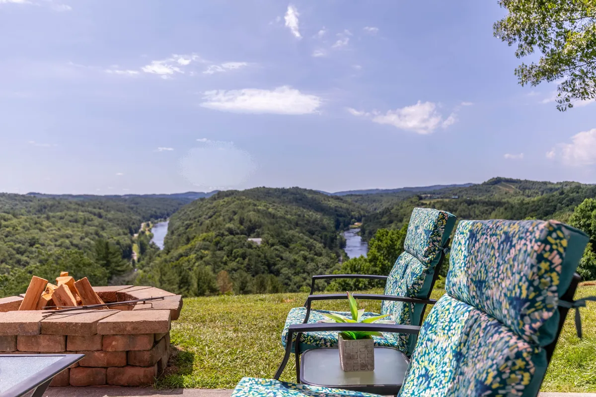 An inviting outdoor fire pit surrounded by lawn chairs overlooks a scenic river and rolling hills. The setting is tranquil, highlighting natural beauty and ample space for relaxation or gatherings with loved ones and pets.