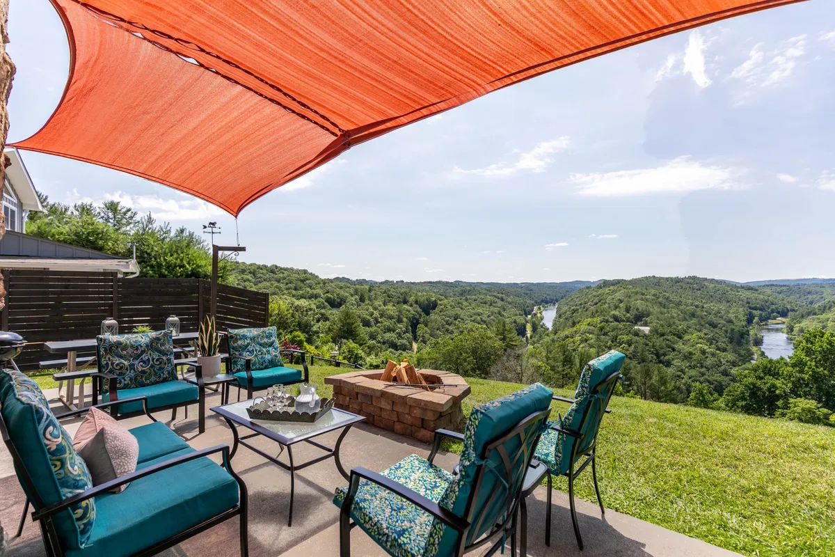 An inviting porch with outdoor seating arranged around a stone fire pit, shaded by a bold orange sail. The porch offers expansive views of the river and lush greenery, making it an ideal space for relaxing, dining, or gathering with friends and family.
