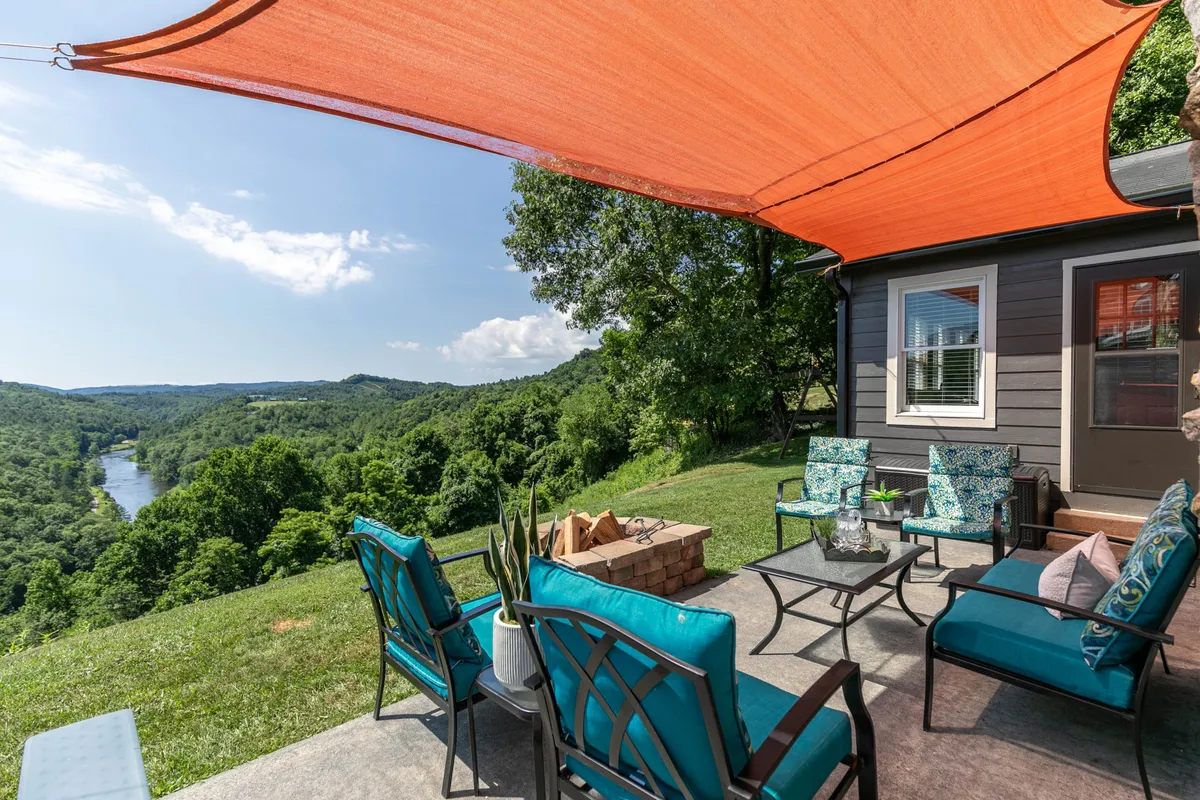 A tranquil outdoor seating area with vibrant blue patio chairs, an orange shade sail for comfort, and a central stone fire pit. The porch overlooks the river, offering stunning views of the surrounding mountains. The scene suggests both relaxation and opportunities for socializing, with the fire pit inviting gatherings at any time of day.