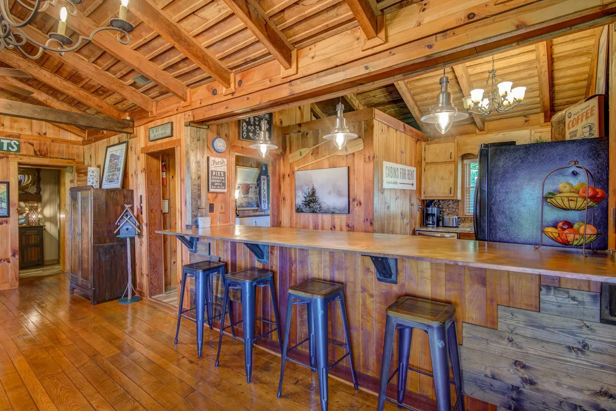 A warm, inviting kitchen scene featuring a wooden bar counter with metal stools, charming wall decor, hanging light fixtures, and a view of a fully equipped kitchen area with modern appliances.